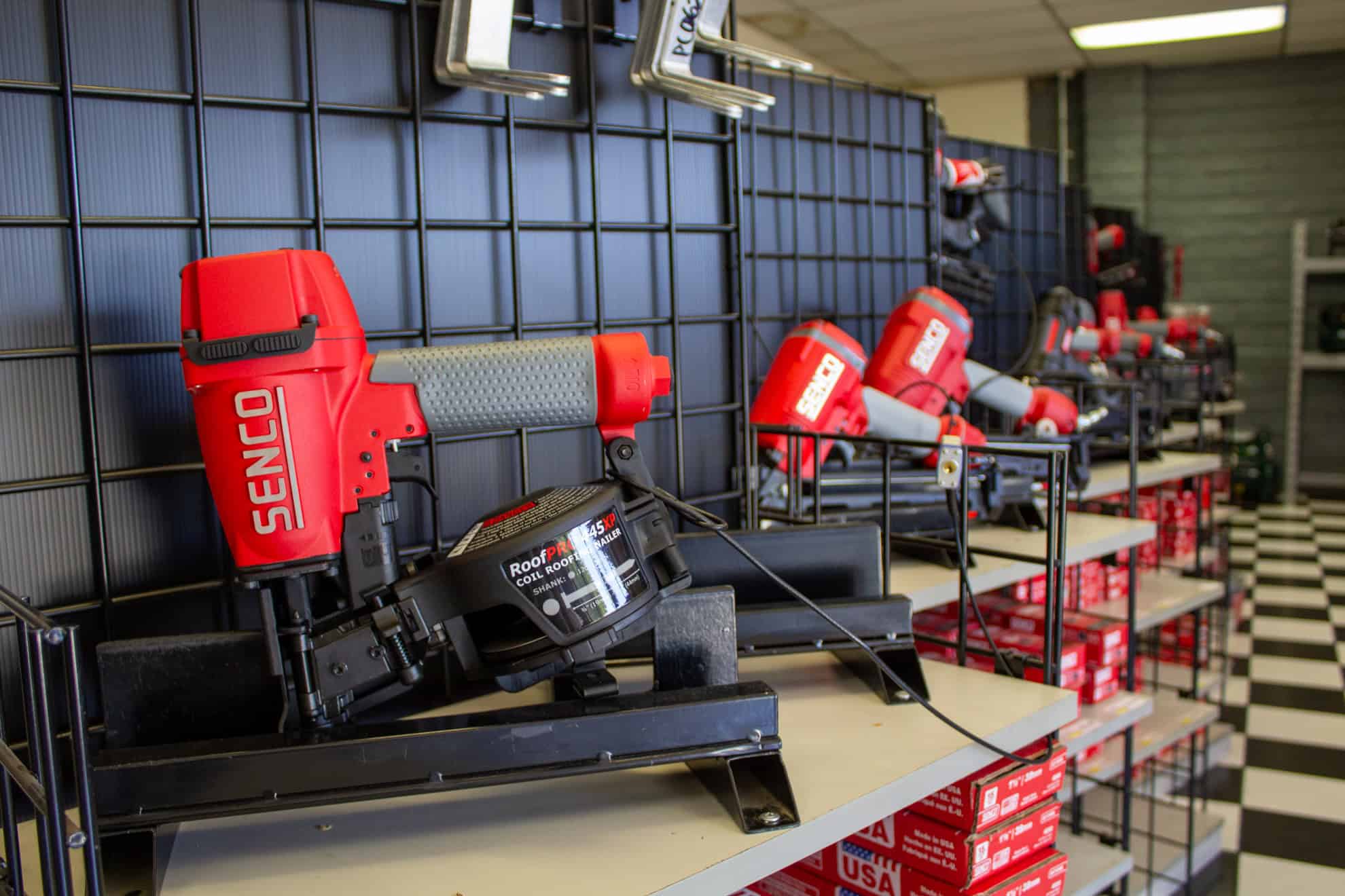 A shelf displays red Senco nail guns and boxes against a black grid wall in a store, with a black and white checkered floor.