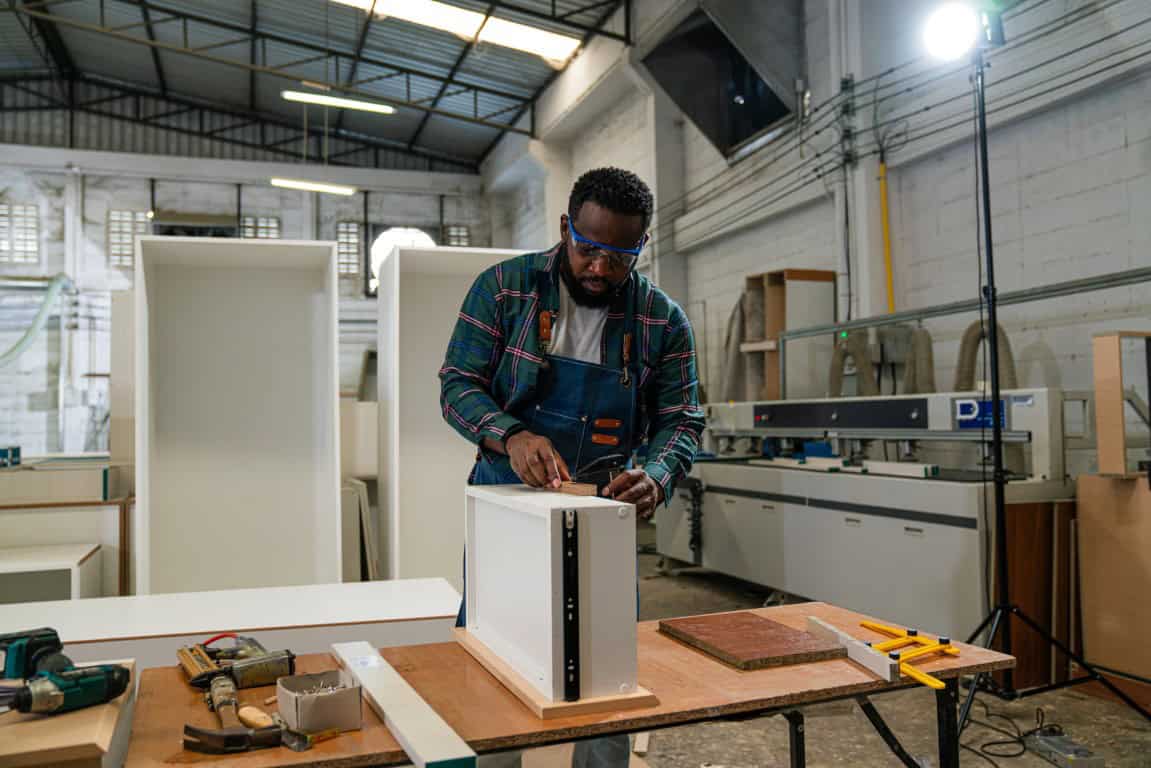 Man wearing safety glasses works on assembling a white cabinet in a workshop, surrounded by tools and woodworking equipment.