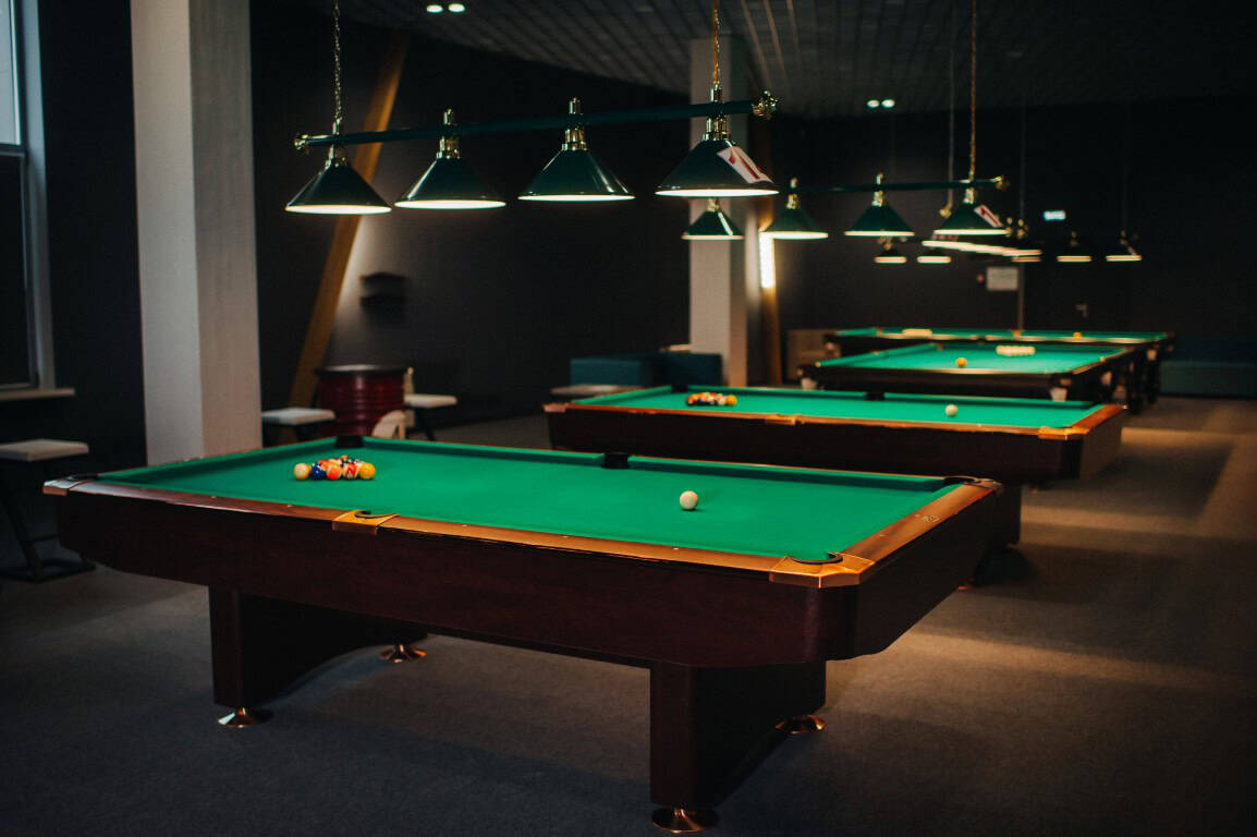 A dimly lit pool hall with multiple green felt pool tables, each set up for a game.