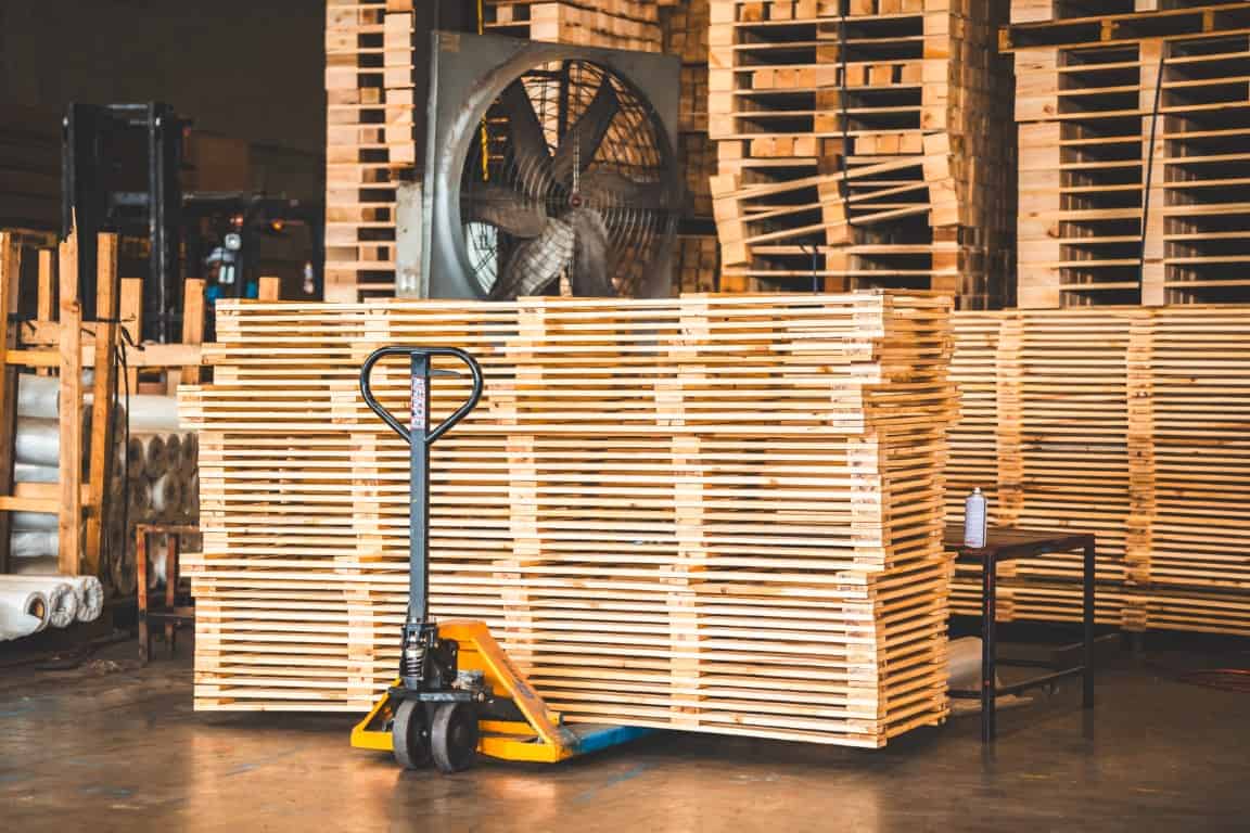 Stacks of wooden pallets in a warehouse, with a pallet jack in front and a large fan in the background.