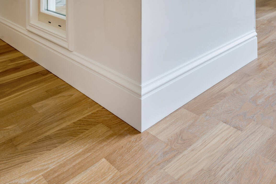 A close-up of a corner in a room shows light wood flooring and white baseboards.