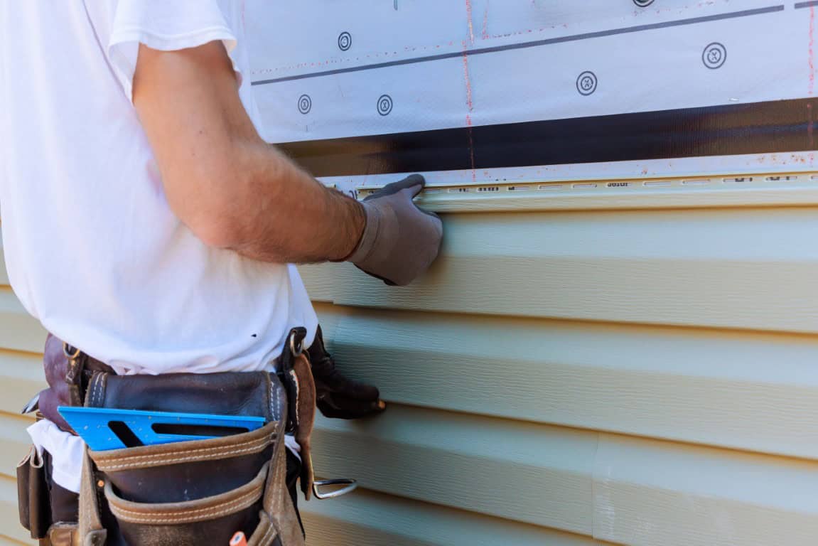 Worker installing green vinyl siding on a building exterior, wearing a tool belt and gloves.