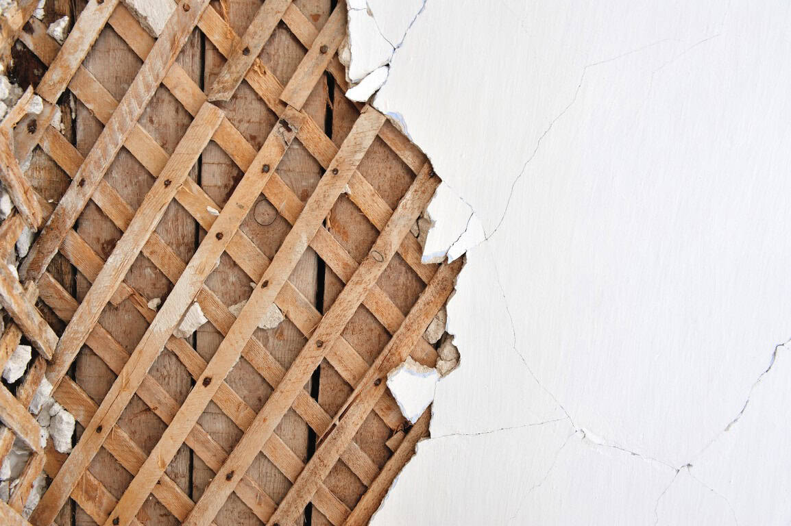 Close-up of a wall with peeling plaster revealing a wooden lath underneath.