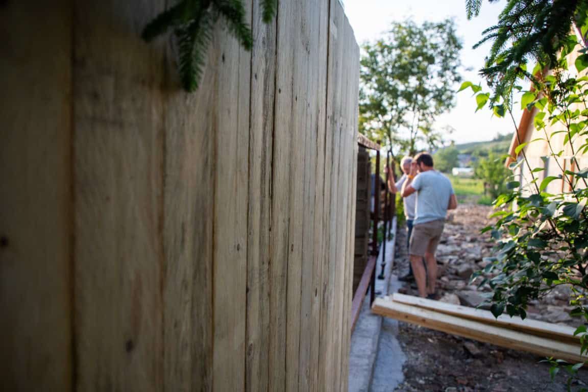 Two people work on constructing a wooden fence in an outdoor setting with trees and a house nearby.