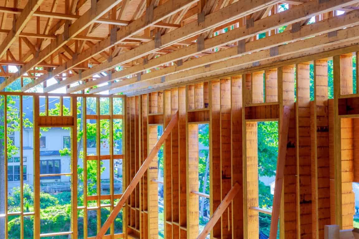 Interior view of a wooden house under construction, showing exposed framing and large window openings, with trees visible outside.