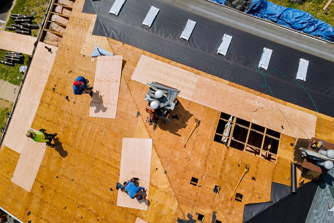 Aerial view of construction workers installing plywood sheets on a roof. Roofing materials and tools are scattered across the structure.