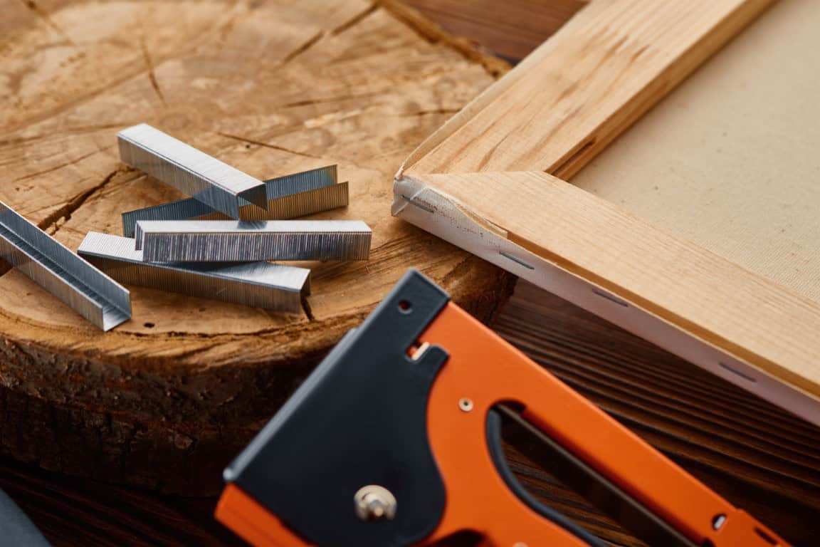 Close-up of a wood frame, a stack of staples, and an orange staple gun on a wooden surface.