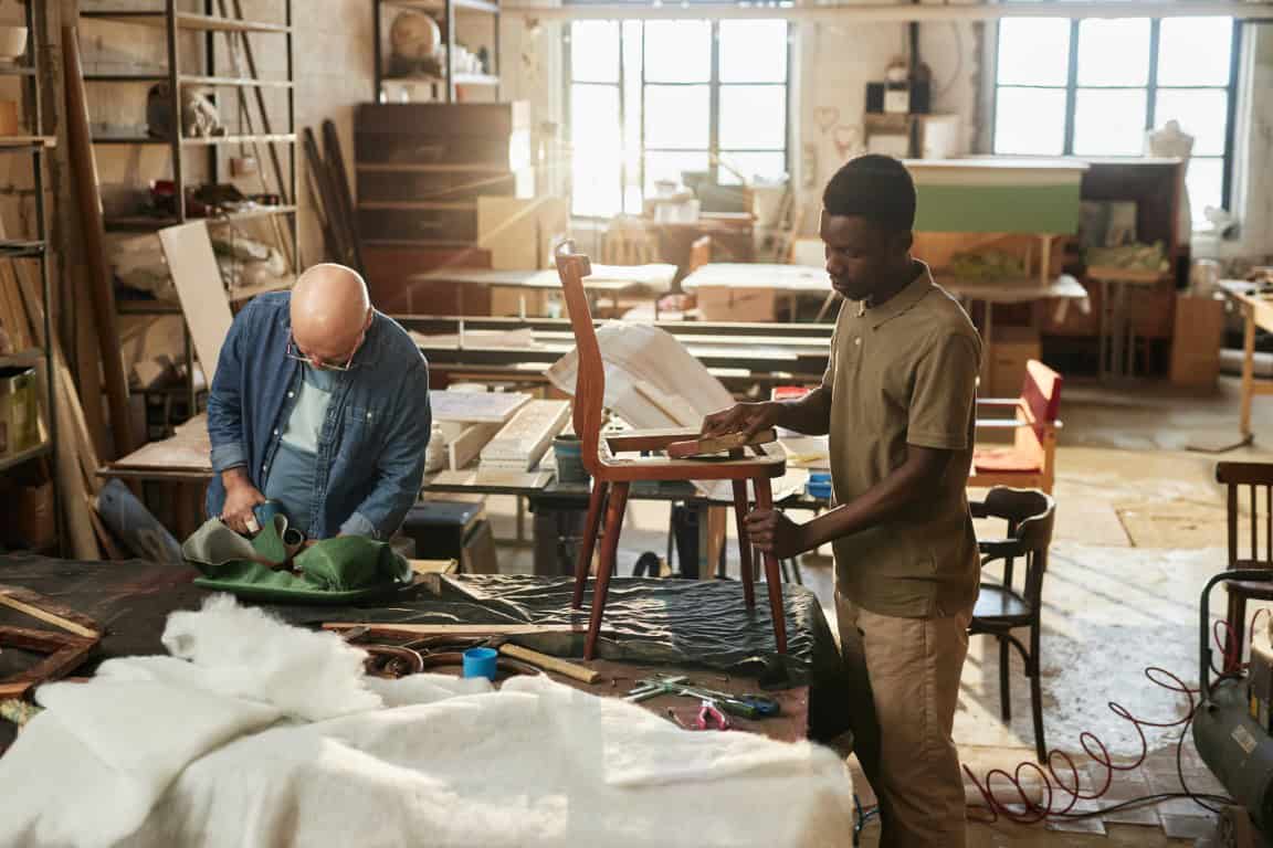 Two men working in a woodworking shop with tools and materials. One man is upholstering a chair, while the other is focusing on a project on the table. Sunlight streams through windows.